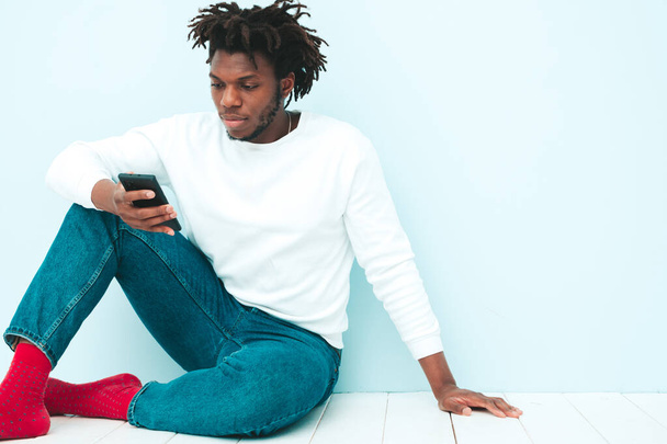 Handsome smiling hipster lambersexual model.Unshaven African man dressed in summer clothes.Fashion male with dreadlocks  posing near blue wall in studio.Looking at smartphone screen.Using apps  - Fotoğraf, Görsel