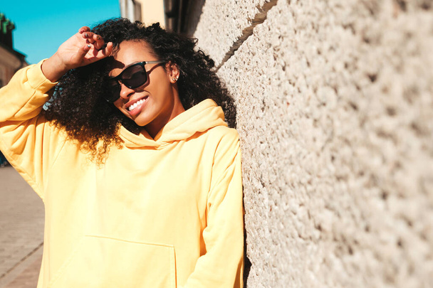 Beautiful black woman with afro curls hairstyle.Smiling hipster model in yellow hoodie. Sexy carefree female posing on the street background in sunglasses. Cheerful and happy - Foto, imagen