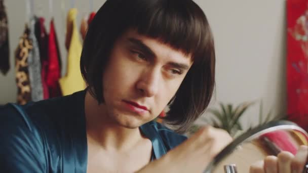 Φωτογραφική μηχανή χειρός του τρανσέξουαλ άνδρα σε μελαχρινή περούκα κρατώντας στρογγυλό καθρέφτη και βούρτσισμα των μαλλιών με χτένα στο σπίτι - Πλάνα, βίντεο