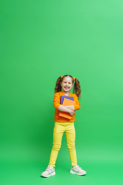 Back to school. a happy little schoolgirl with two ponytails, in fashionable clothes with books in her hands. a little girl smiles to prepare for school and poses on a green background. - Photo, image