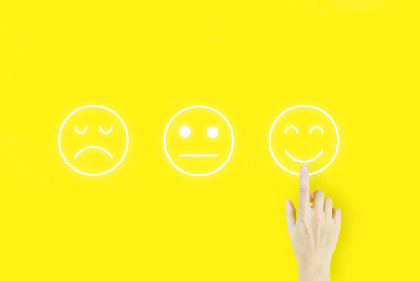 Customer service evaluation and satisfaction survey concepts. Young woman's hand finger pointing with hologram face emotion on yellow background - Photo, Image
