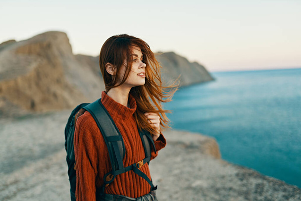 happy traveler in a red sweater with a backpack on her back look at the sea in the distance and high mountains - Photo, Image