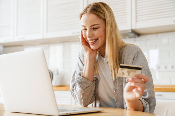 Joyful blonde girl using laptop and credit card while sitting at home kitchen - Фото, изображение