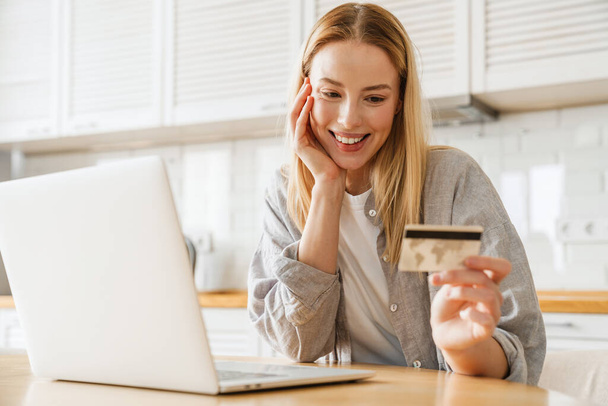 Joyful blonde girl using laptop and credit card while sitting at home kitchen - Foto, Imagem