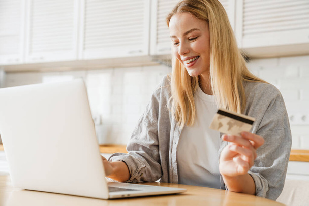 Joyful blonde girl using laptop and credit card while sitting at home kitchen - Photo, image