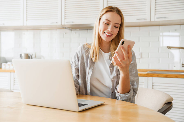 Radosna blondynka za pomocą laptopa i smartfona podczas siedzenia w domu kuchnia - Zdjęcie, obraz