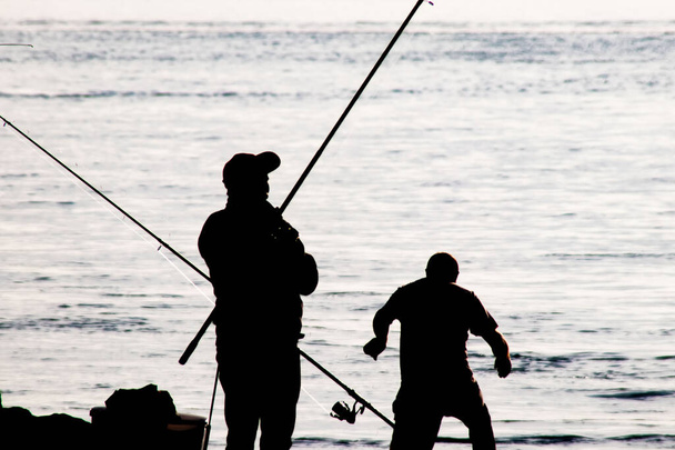 Siluetas de pescadores aficionados con cañas de pescar en la orilla del mar temprano en la mañana. - Vector, imagen