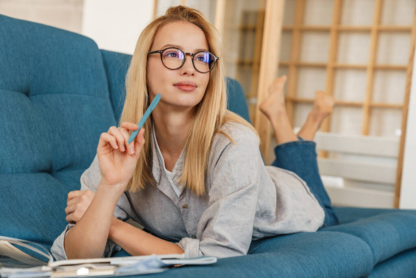 Thinking student girl in eyeglasses doing homework while lying on couch at home - Foto, Imagen