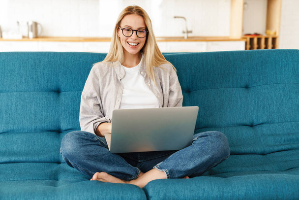 Attractive young smiling woman using laptop computer while sitting on a couch at home - Foto, immagini
