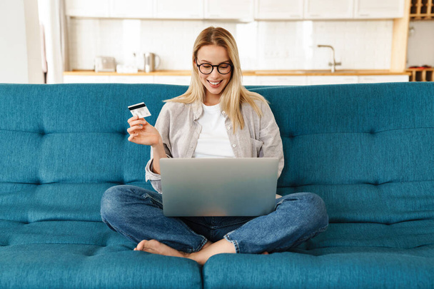 Smiling attractive young woman shopping online via laptop computer and credit card while relaxing on a couch at home - Φωτογραφία, εικόνα