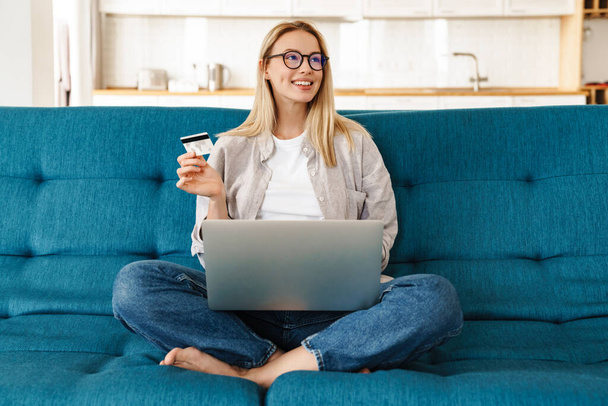 Smiling attractive young woman shopping online via laptop computer and credit card while relaxing on a couch at home - Photo, image