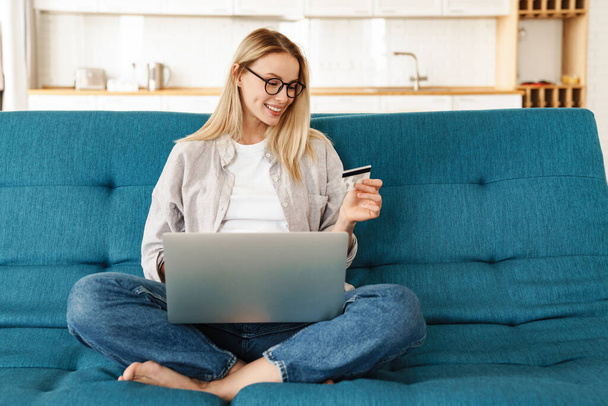 Smiling attractive young woman shopping online via laptop computer and credit card while relaxing on a couch at home - Photo, Image