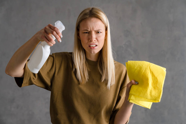 Displeased european woman posing with cleaning spray and rag isolated over grey wall - Foto, Imagem