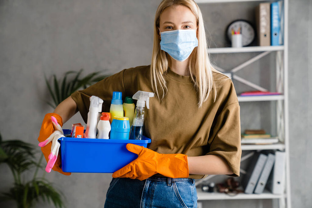 Blonde european woman in face mask posing with cleaning product at home - Φωτογραφία, εικόνα