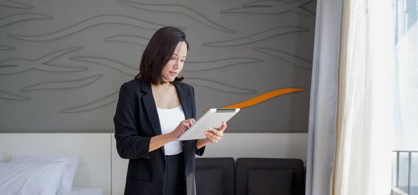 Ασιάτισσα επιχειρηματίας με μαύρο κοστούμι στέκεται με tablet υπολογιστή στο χέρι της. Ατμόσφαιρα της εργασίας έξω από το γραφείο κατά τη διάρκεια των διακοπών. - Φωτογραφία, εικόνα