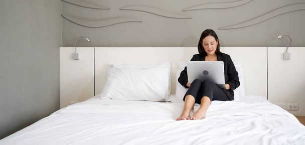 Азиатка в черном костюме использует ноутбук, чтобы работать утром в постели. - Фото, изображение