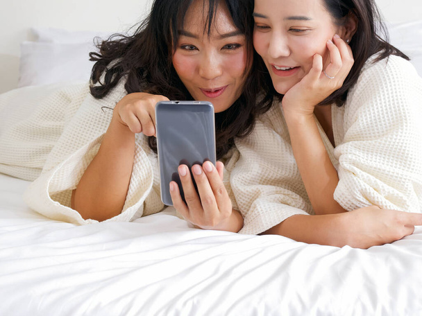 Two Asian women in bathrobes take a selfie with a mobile phone while lying in bed. - Fotoğraf, Görsel