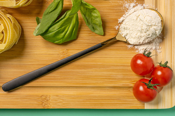 Top view image of spoon with flour, cherry tomatoes, basil and bamboo table on plain green background - Photo, Image