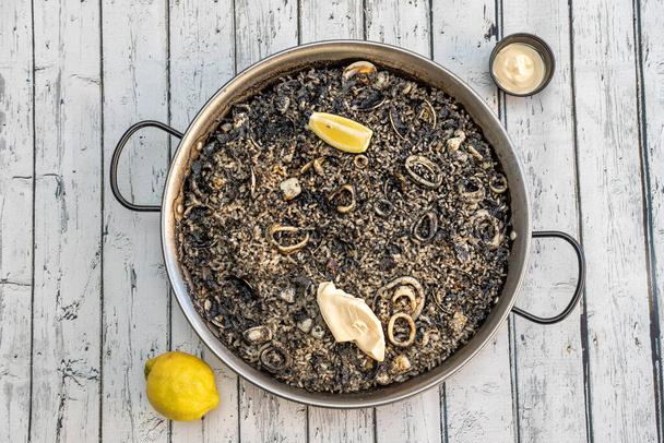 Paella española de arroz negro con calamares, pescado y almejas con salsa aioli y limón sobre mesa blanca - Foto, Imagen