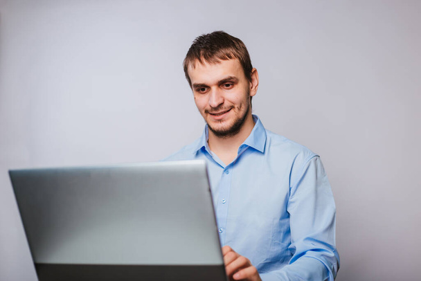 青いシャツを着た若いハンサムな男がコンピュータに座っている。白い背景のオフィスの労働者はノートパソコンのために働く。隔離 - 写真・画像