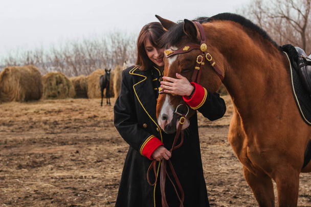 Kaunis nainen, jolla on pitkät ja mustat hiukset historiallisessa husaarin puvussa, seisoo pellolla hevosen kanssa. Tyttö, jolla on hevonen - Valokuva, kuva