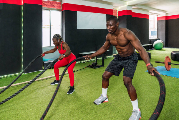 Two black bodybuilder athletes in a sports gym doing an aerobic rope exercise with a lot of effort to strengthen arms and chest. Concept of strengthening the body in gym - Photo, Image