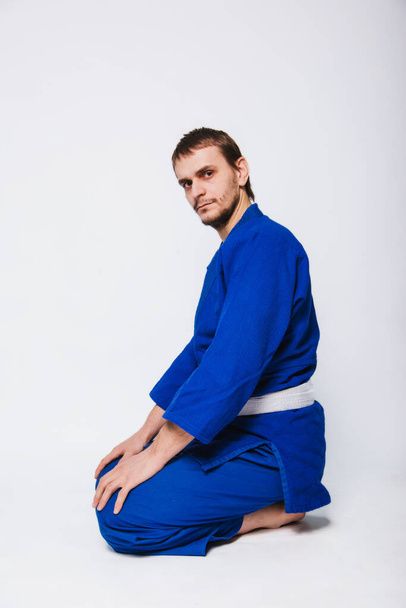 Een man in een blauwe kimono met een witte sjerp zit op de vloer. Een beginner vechter van oosterse vechtsporten - Foto, afbeelding