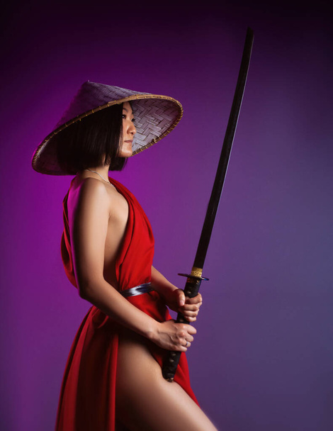 nahá štíhlá asijská žena v červené pláštěnce a asijský klobouk s katanou v její ruce obraz samuraje na neonovém pozadí - Fotografie, Obrázek