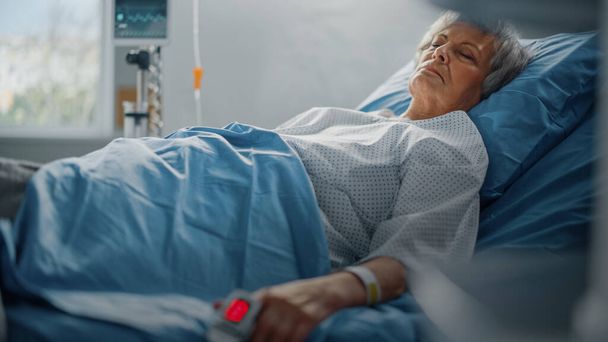 Hospital Ward: Senior Woman Descansando en una cama con Finger Heart Rate Monitor Oxímetro de pulso que muestra Pulso. Sus manos frágiles descansando sobre una manta. Concéntrate en la mano. - Foto, imagen