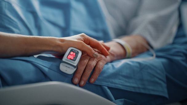 Hospital Ward: Senior Woman Resting in a bed with Finger Heart Rate Monitor, Caring Family Member Holds Her Fragile Hand, Support and Comforting. Focus on the Hands. - Фото, изображение