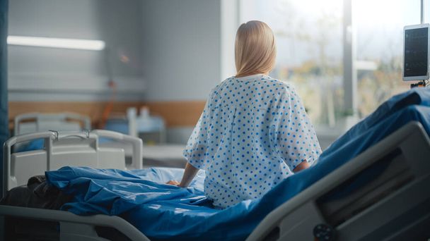 Hospital Ward: Beautiful Caucasian Female Patient Sitting on a Bed, Fully Recovering after Successful Surgery, Covid-19, Sickness she Looks Around and out of the Window with a Beautiful Sunny View - Фото, изображение