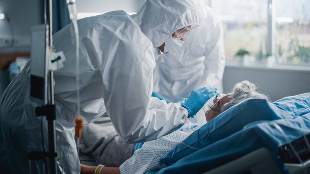 Hospital Coronavirus Emergency Department Ward: Team of Doctors wearing Coveralls, Face Masks Take Care of a Senior Patient Lying in Bed, Put Oxygen Mask and Lung Ventilator. Medics Saving Lives - Фото, зображення