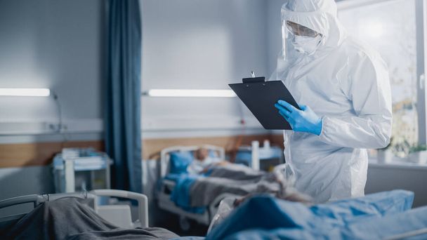 Hospital Coronavirus Emergency Department Ward: Doctor wearing Coveralls, Face Mask Take Care of a Senior Patient Lying in Bed, Talks with Patient, Fill Medical Data. Medics Saving Lives - Фото, изображение