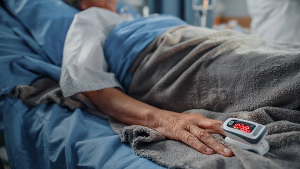 Hospital Ward: Senior Woman Resting in a bed with Finger Heart Rate Monitor Pulse oximeter showing Pulse. Her Fragile Hands Resting on a Blanket. Focus on the Hand. - Foto, Imagem