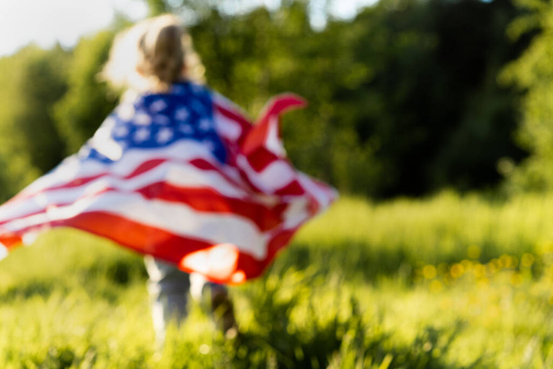 piękna mała blondynka z amerykańską flagą w przyrodzie w świetle słonecznym, zdjęcia w pełnym skupieniu. - Zdjęcie, obraz