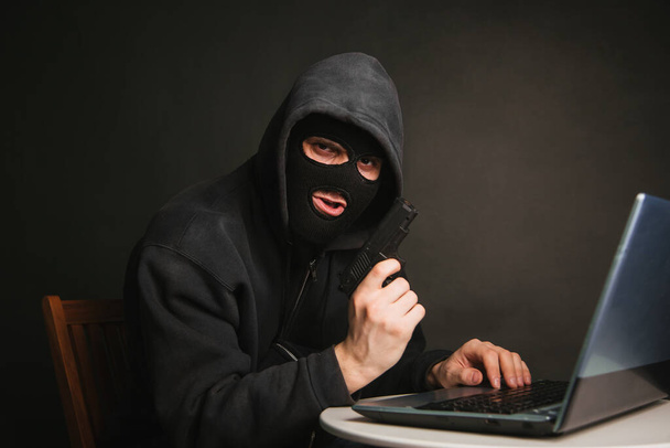 Een crimineel met een zwart masker en een kap met een pistool steelt geld via een laptop. Hacker aanvallen. Onlinediefstalconcept - Foto, afbeelding