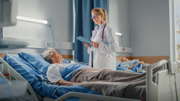 Hospital Ward: Friendly Doctor Asks Health Care Questions of Smiling Elderly Lady Resting in Bed. Physician Uses Tablet Computer, Does Checkup, Talk to Old Woman Fully Recovering after Sickness - Foto, imagen