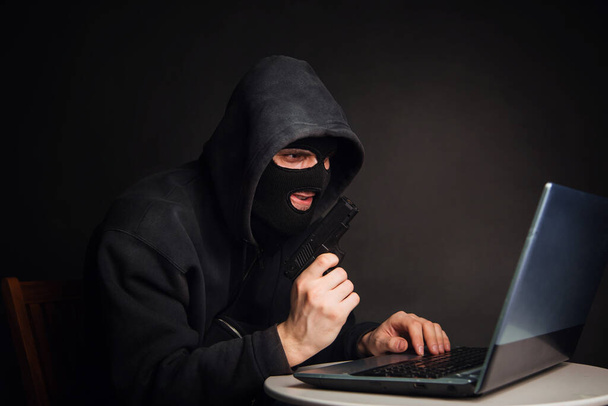 Przestępca w czarnej masce i kapturze z pistoletem kradnie pieniądze przez laptopa. Ataki hakerów. Koncepcja kradzieży online - Zdjęcie, obraz