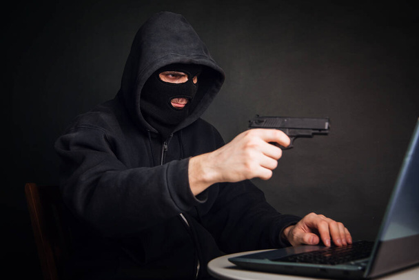黒いマスクの犯罪者とピストルを持ったフードはノートパソコンを通してお金を盗む。ハッカーの攻撃だ。オンライン盗難の概念 - 写真・画像