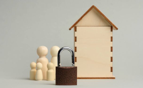 Metallschloss auf dem Hintergrund eines hölzernen Miniaturhauses und Holzfiguren einer Familie auf grauem Hintergrund. Sicherheitskonzept für Daten und Privateigentum, Sicherheitssystem - Foto, Bild