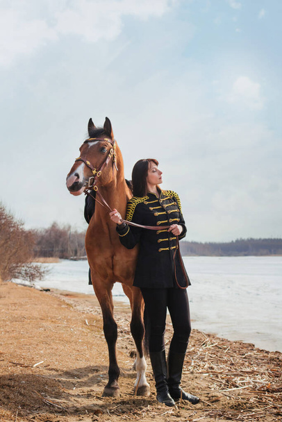 Kaunis nainen, jolla on pitkät ja mustat hiukset historiallisessa husaarin puvussa, seisoo lähellä jokea hevosen kanssa. Tytöllä hevonen - Valokuva, kuva