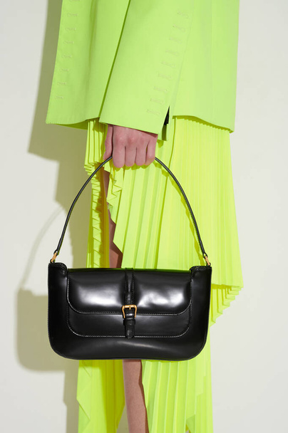 Chica en chaqueta verde ácido y falda plisada con bolso de cuero negro de lujo en la mano. Foto de moda vertical. Vista lateral. - Foto, Imagen