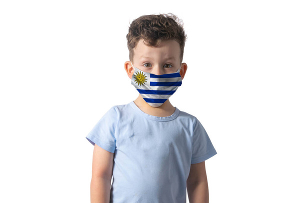 Atemschutzmaske mit Fahne von Uruguay. Weißer Junge setzt medizinische Gesichtsmaske isoliert auf weißem Hintergrund auf. - Foto, Bild