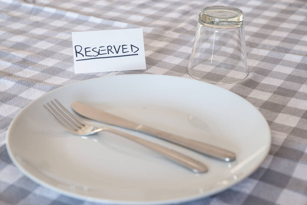 Knife, fork, glass, with card with "reserved" text, on gray checked tablecloth. Booking a table in a restaurant.  - Photo, Image