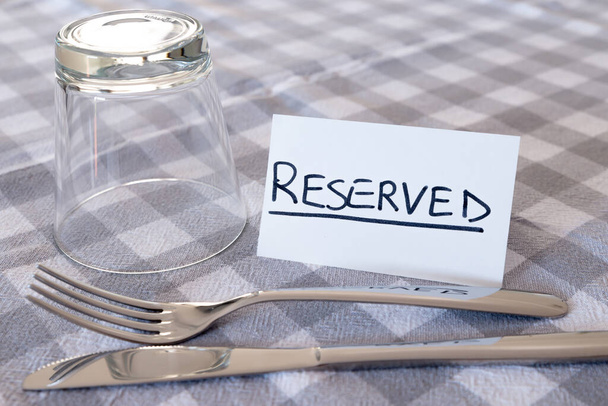 Messer, Gabel, Glas, mit Karte mit "reserviertem" Text, auf grau karierter Tischdecke. Tischreservierung im Restaurant.  - Foto, Bild