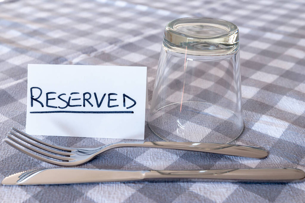 Knife, fork, glass, with card with "reserved" text, on gray checked tablecloth. Booking a table in a restaurant.  - Photo, Image