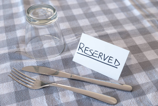 Faca, garfo, vidro, com cartão com texto "reservado", em toalha de mesa cinza verificada. Reserva de uma mesa em um restaurante.  - Foto, Imagem