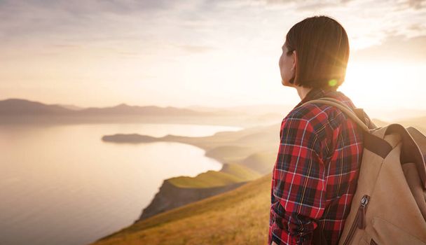 A young Asian woman with a backpack hiking in the summer and enjoying the view of the sunset sea and mountains. Mountain and coastal travel, freedom and an active lifestyle - Photo, Image