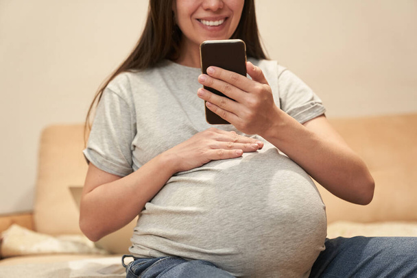 Terhes nő otthon telefonnal ül és mosolyog, videokommunikáció - Fotó, kép