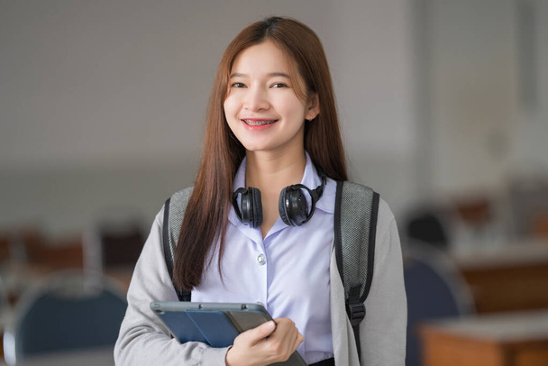 Portret młodej azjatyckiej studentki w mundurze trzymającej tablet w eleganckiej i szczęśliwej pozie w klasie uniwersyteckiej lub na studiach. Młodzież uczennica i korepetycje edukacja z koncepcji uczenia się technologii. - Zdjęcie, obraz
